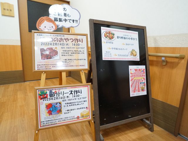 案内掲示物と廊下 展示物 明るい廊下に立つ案内掲示物には、様々なイベントの告知がなされており、利用者の交流を促しています。