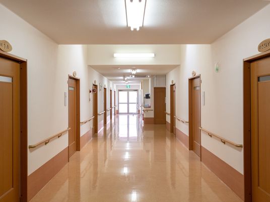 施設の写真 通路幅のある廊下。落ち着いたブラウン色のフローリングで壁紙は白い。両サイドに木製の手すりが設置されている。