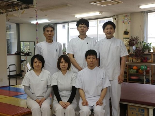 スタッフ集合の多目的室 多目的室   白い制服のスタッフが複数人並び、明るい多目的室で落ち着いた雰囲気です。運動用のマットや備品も見え、働く場の様子が伝わります。