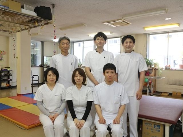 スタッフ集合の機能訓練室 機能訓練室   白衣のスタッフが集まり、明るい機能訓練室で撮影されています。運動用マットや器具が見え、支援の現場をイメージしやすいです。