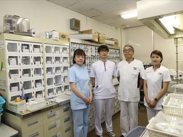 薬剤管理室のスタッフ 内観   整理された収納棚が並ぶ薬剤管理室で、4人のスタッフの方が並んで立っています。白衣姿の様子から、落ち着いて連携しやすい環境が伝わります。