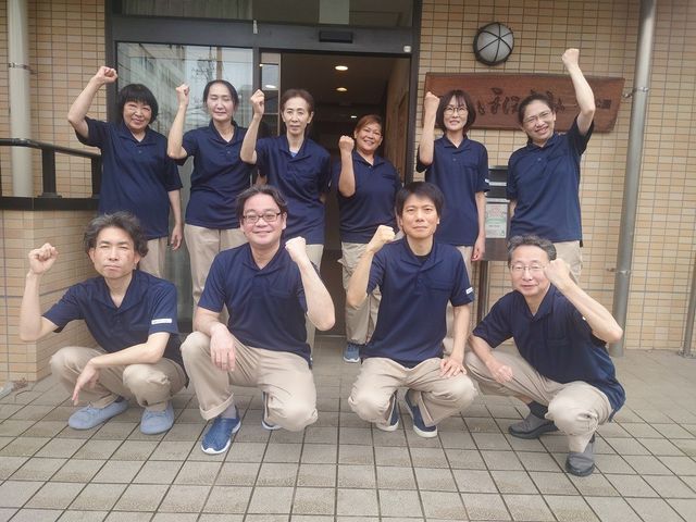 スタッフ集合のエントランス エントランス   紺色の制服でそろえたスタッフが、入口前で笑顔と元気なポーズを見せています。職場の雰囲気が伝わり、チームで支える様子が感じられます。