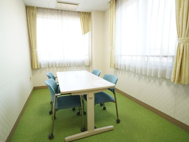 緑のカーペットの会議室 共有スペース ゆったりとした空間に配置されたテーブルと椅子が、明るい窓辺で利用者の交流を支えます。白光が射し込むこの部屋は、皆様のコミュニケーションの場として最適です。
