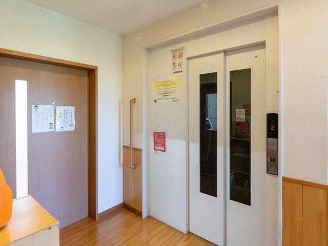 明るいエレベーター前 廊下   木目調の床とやわらかな照明で、落ち着いた雰囲気のエレベーター前です。壁面の掲示も見やすく、動線の確認がしやすそうです。