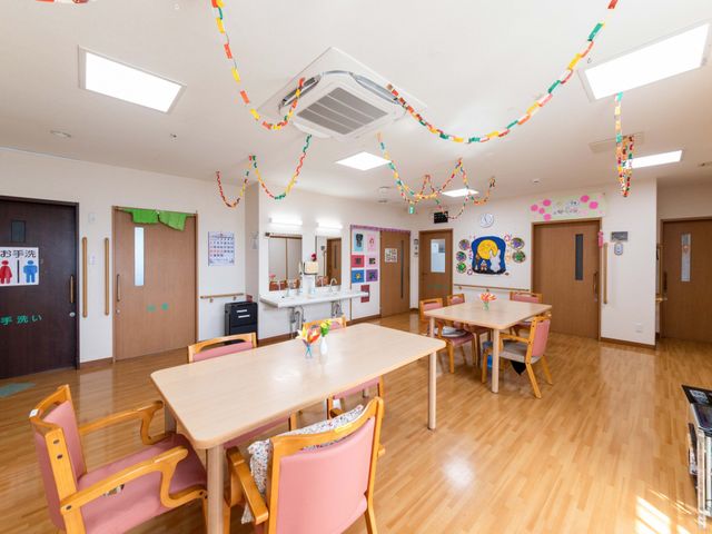 明るい食堂の様子 食堂 木目調の床と明るい照明で、落ち着いて過ごしやすい雰囲気の食堂です。テーブル席がゆったり配置され、手洗い設備も見えます。
