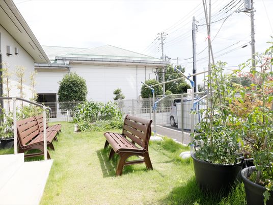 施設の建物の前に芝生の庭がある。背もたれのある長いベンチが３脚置かれている。トマトの植えられたプランターがあり、奥には菜園が見える。