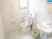サムネイル 壁、床ともに白で統一され、清潔感のあるトイレである。温水洗浄機能付き便座が設置されている。隣には洗面台が設けてある。