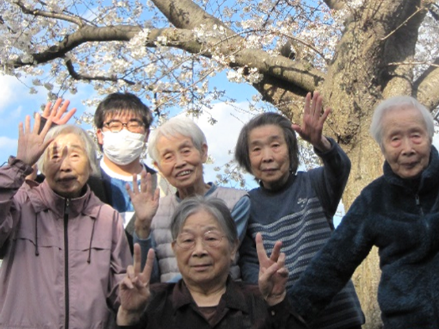 桜の下で笑顔の集合 外出 桜が咲く清々しい屋外で、利用者同士が親交を深める様子です。新たな職場での新鮮な出会いが待っています。