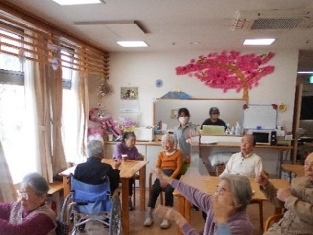 日中の明るい共有スペース リビング ゆったりとした空間で行われるレクリエーションの様子です。笑顔で健康体操をする利用者の方々を見守るスタッフも写っています。