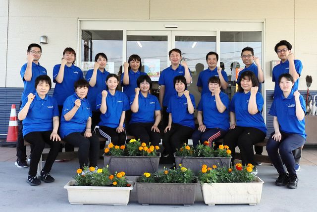 スタッフ集合の笑顔 元気で明るい印象のスタッフが、チームワークを感じさせるポーズで団結しています。職場の雰囲気としては、協力的で和やかな環境がうかがえます。
