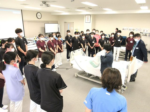機能訓練の説明場面 機能訓練室   広い訓練スペースで、ベッドを使いながらスタッフが説明しています。多くの職員が集まり、実践的な学びの場になっています。