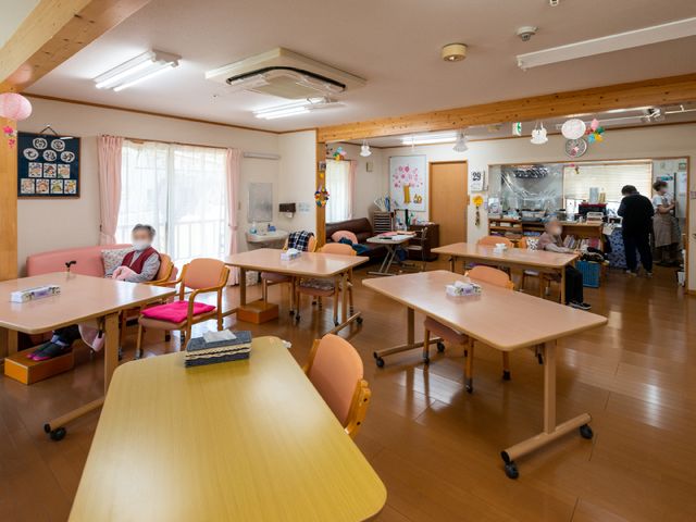 座って過ごす居住者の食堂 共用スペース 天井の高い食堂で、居住者が心地よいひとときを過ごしています。明るく開放的なこの空間でのお食事や交流が、日々の楽しみの一つになります。