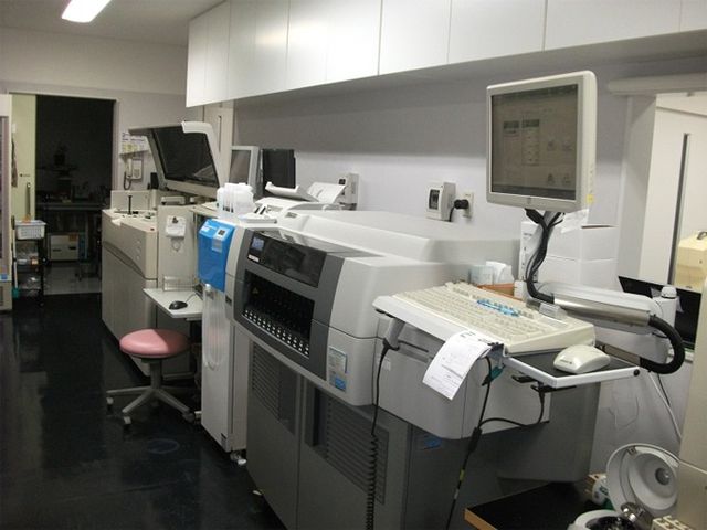 検査機器が並ぶ室内 内観   検査機器やモニターが並ぶ、清潔感のある作業スペースです。奥には別室へ続く出入口も見え、落ち着いて業務に取り組めそうです。