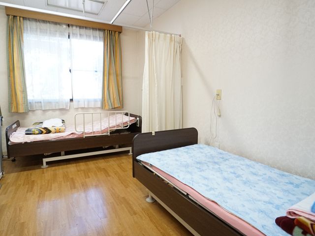 ゆったり居室のベッド 居室 ゆとりのある居室には、日当たりの良い窓と清潔感のあるベッドが備え付けられ、安らぎの空間を提供しています。