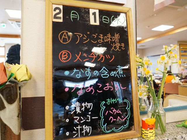 掲示板と花がある空間 展示物 明るい空間での昼食メニューが書かれたボード。日々の健康的な食事を提供する環境です。