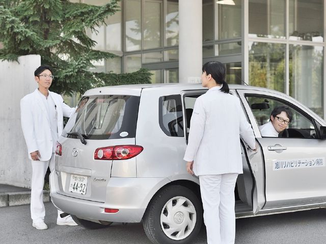 送迎車の前で職員が対応する外観 外観   送迎車のそばで職員の方が声をかけ合っています。出入り口付近での連携が見え、就業時の動きがイメージしやすいです。