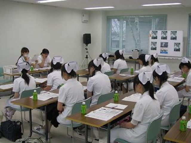 看護学生が学ぶ多目的室 多目的室   机を並べた明るい多目的室で、看護服の方々が着席して学んでいます。就業前の研修や勉強会にも使いやすそうな空間です。