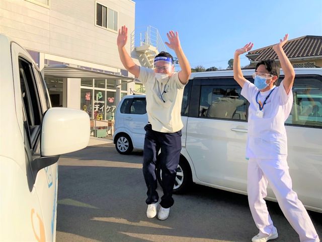 送迎中のスタッフと車両の外観 外観   施設前でスタッフの方が車両のそばに立ち、元気に手を上げています。送迎対応の様子が見え、働く場面をイメージしやすいです。