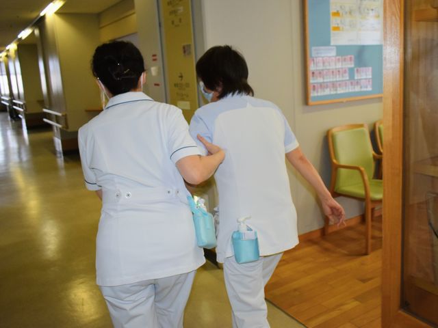 スタッフが廊下を歩く様子 廊下   スタッフ同士が声をかけ合いながら廊下を移動しています。手すりや待合用の椅子があり、見守りしやすい環境です。