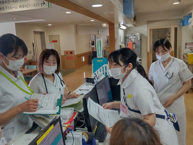 白衣のスタッフが集まる廊下 廊下   白衣のスタッフが複数人で書類を確認しています。広い通路に案内表示があり、連携しながら動きやすい印象です。