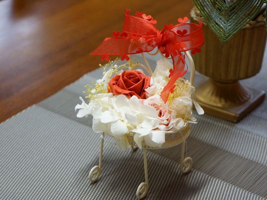 木目調デザインのテーブルの上に、上品なライトグレーの敷物が置いてある。その上に、赤いリボンの付いた花の装飾品がある。
