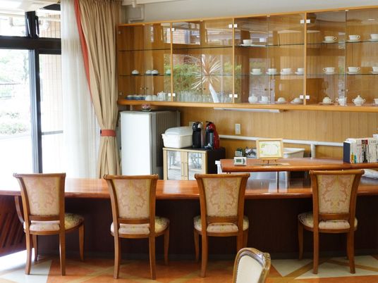 ライトブラウンのテーブルの周りに、４脚の椅子がセットされている。奥には大きな食器棚があり、数多くのコーヒーカップが並べられている。