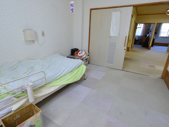 居室の広いスペース 居室のドア 広々とした居室には、ベッドと個人のスペースが確保されており、自分の時間を大切に過ごせる環境が整っています。遠くには共有のリビングが見え、そこでの交流も期待できます。