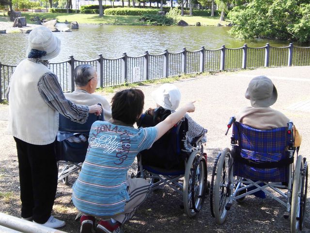 池のそばのリラックス 庭 穏やかな池のそばで、車椅子の方々が介護士と共にリラックスしています。自然に囲まれた静かな環境です。