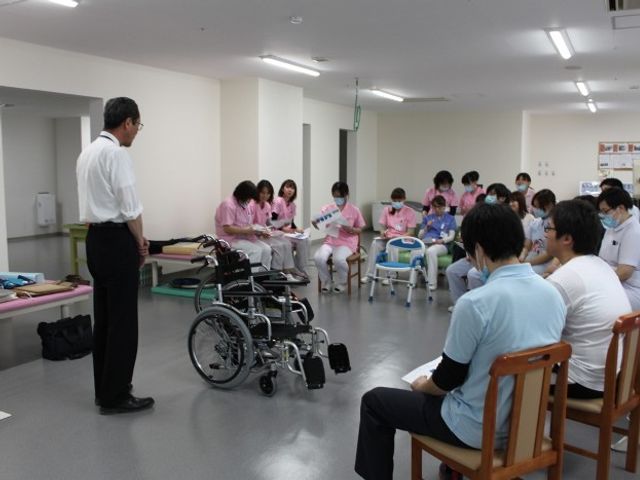 機能訓練室での説明 機能訓練室   広い室内に車椅子や歩行器が並び、職員が利用者へ説明しています。研修や訓練の様子が見え、働く場の雰囲気が伝わります。