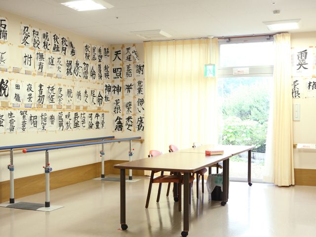 書道作品が掲示された部屋 部屋 壁に掲げられた書道の作品が、心温まる雰囲気を作り出しており、ここでの生活が充実していることが伺えます。明るい窓からは自然の光が差し込んでおり、開放感のある空間です。