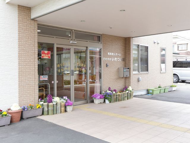 エントランスの花々と施設 出入り口 暖かい歓迎の雰囲気が漂うエントランスです。ここから新たなお仕事が始まりますので、ぜひ一緒に働きましょう。