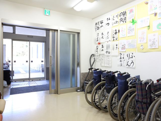 エントランスと掲示物 玄関 壁には色とりどりの作品や掲示物が飾られ、訪れる人を温かく迎えてくれます。車椅子も整然と並べられ、誰もが安心して出入りできるよう配慮されています。