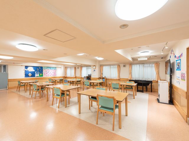 広々としたダイニングルーム 食堂兼機能訓練スペース 広々とした明るい食堂で、皆様が集い交流を楽しむ場所です。天井の高い開放的な空間で、ここでの時間を彩るアクティビティも豊富にご用意しています。