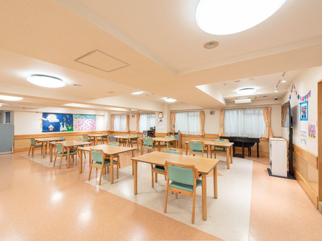 広々としたダイニングルーム 食堂兼機能訓練スペース 広々とした明るい食堂で、皆様が集い交流を楽しむ場所です。天井の高い開放的な空間で、ここでの時間を彩るアクティビティも豊富にご用意しています。
