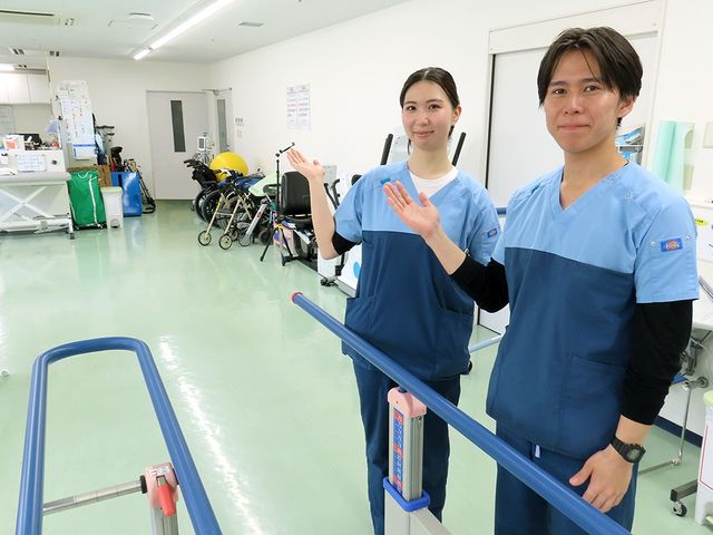 機能訓練室のスタッフ 機能訓練室   明るく広い訓練スペースで、スタッフの方が笑顔で案内しています。歩行練習用の手すりや車椅子があり、支援の様子が伝わります。