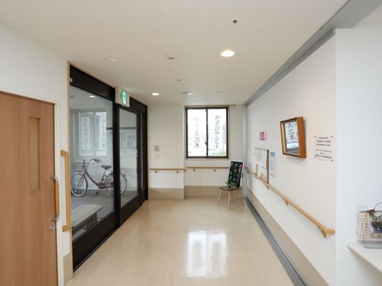 施設の写真 ベージュ色の床は、段差のないバリアフリー構造である。壁際に椅子が置いてあり、黒い看板と観葉植物が飾ってある。