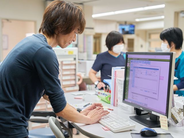 スタッフが確認する受付 フロント   スタッフがパソコン画面を見ながら業務を進めています。明るい受付で、複数の職員が連携している様子が伝わります。