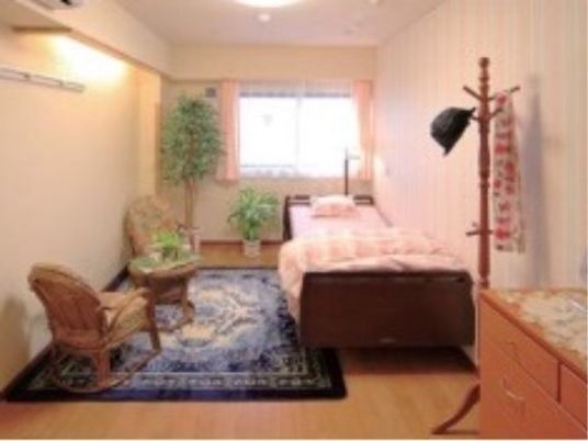 白を基調とした明るい居室である。介護用ベッドやチェスト、ハンガー、椅子、テーブルなど生活に必要な備品がそろっている。