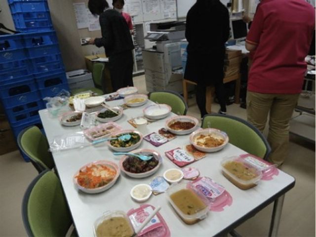 多様な料理が並ぶ食堂 お食事２ 手作り感のあふれるバラエティ豊かな料理が並び、温かな雰囲気の職場です。スタッフ間のコミュニケーションも活発そうで、明るい職場環境を感じ取れます。