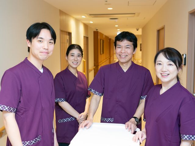 笑顔のスタッフが並ぶ廊下 廊下   明るい廊下で、4人のスタッフが笑顔で並んでいます。木目調の扉や手すりが見え、落ち着いた雰囲気です。