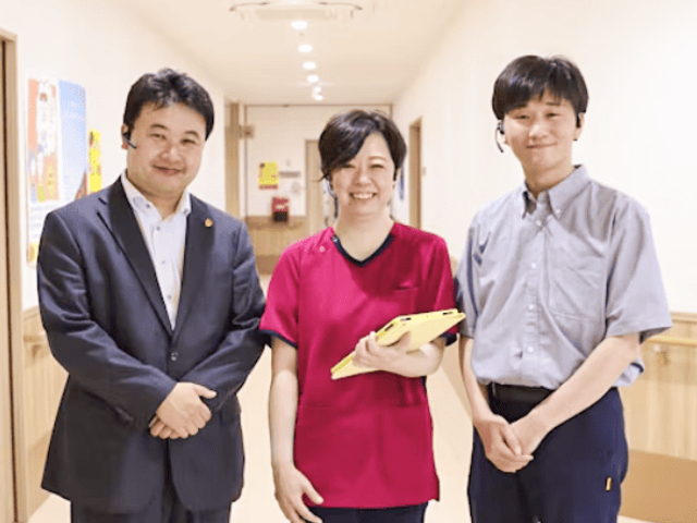 廊下に立つ職員たち 廊下   明るい廊下で、3人の職員の方が並んでいます。清潔感のある通路で、連携しやすい雰囲気が伝わります。
