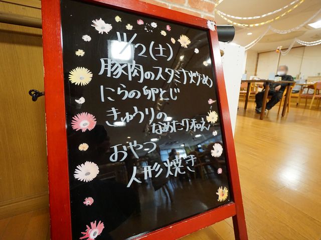 スケジュール掲示板 展示物 手書きで配慮された案内板が食堂入口に置かれ、安らぎの時間を過ごせる空間を伝えています。お客様がくつろぐ姿も見え、温かな雰囲気が感じられます。