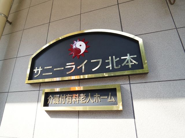 看板のロゴと名称 ロゴ 落ち着いた色合いの看板が目を引く明るい場所です。来訪者や働く皆さまを温かく迎えるエントランスの一部であることが伝わります。