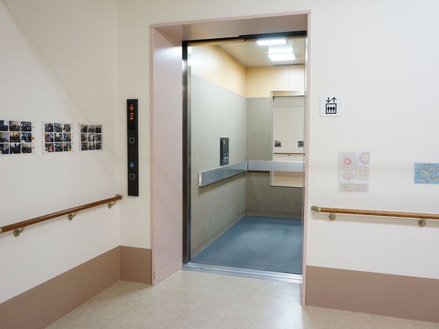 広々とした廊下のエレベーター エベーター 手すりが整った廊下に、車椅子でも利用しやすい広いエレベーターが設置されております。壁には暖かみのあるアートが飾られ、快適な移動が期待できます。