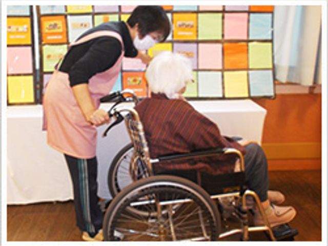 会話中の様子の居室 居室 介護士が車椅子の利用者と一緒に掲示物を見ています。温かみのある和やかな雰囲気です。