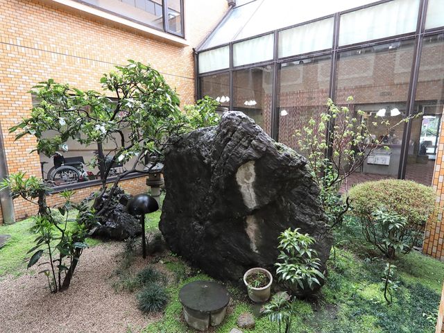 和風の緑豊かな中庭 日本庭園 石と緑に囲まれた落ち着いた雰囲気の中庭です。車椅子で外を眺める方の姿から、ゆっくりとした時間が流れる空間が感じられます。