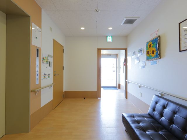 廊下の手摺りとソファー エレベーター前の廊下 明るい廊下には、安全に配慮した手すりが備えられ、壁には温かみのある装飾が施されています。ソファーでの休憩もでき、訪れる方々に心地よい空間を提供しています。