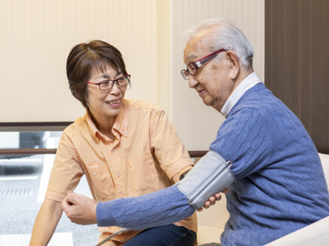 血圧測定をする利用者と職員 職員が笑顔で利用者の健康管理を行う様子が伺えます。ここでは心のこもった支援が日々実施されています。