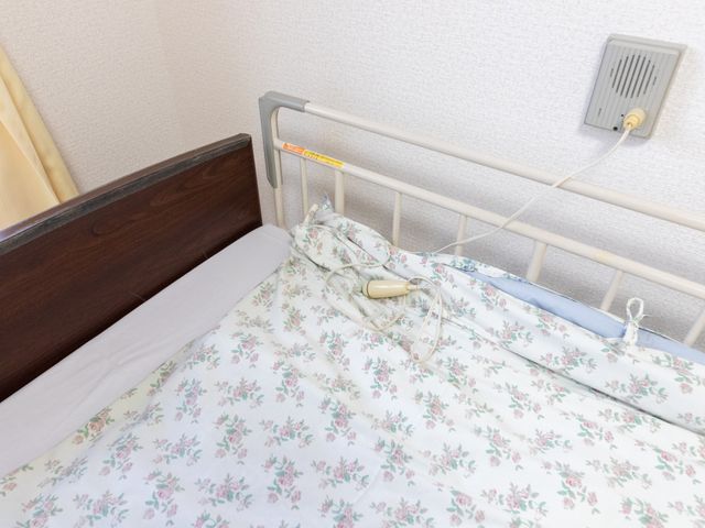 花柄寝具の居室 居室   明るい壁紙と落ち着いた木目のヘッドボードが印象的な居室です。ベッドまわりには手すりやナースコールがあり、安心して過ごしやすそうです。