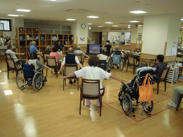 車椅子の人々が運動する室内 機能訓練室 明るい室内では、車椅子の方も含めて皆で楽しく体操が行えます。交流の場としても利用できます。